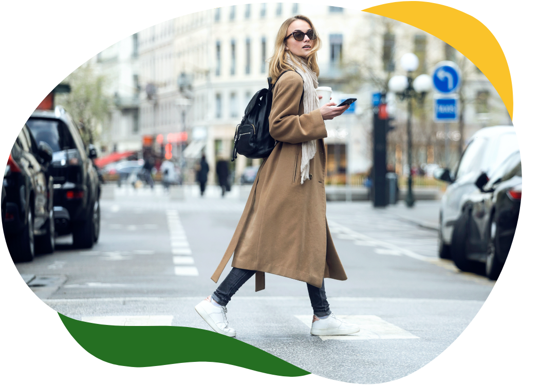 Moderná žena s veľkými slnečnými okuliarmi v dlhom béžovom kabáte prechádza cez cestu a v pravej ruke drží mobil