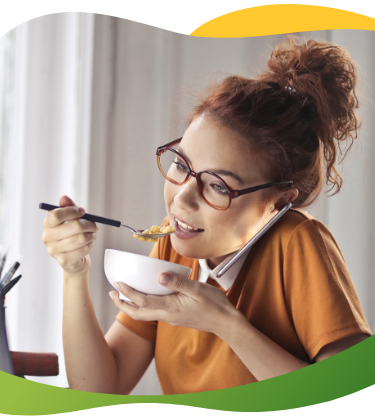 Mladá žena v okuliaroch s vyčesanými vlasmi sedí pred notebookom, telefonuje a súčasne konzumuje jedlo